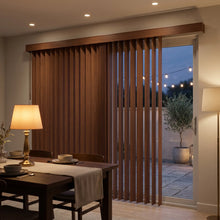 Load image into Gallery viewer, Warm and inviting Wood Grain Faux Wood PVC Vertical Blinds in Brown perfectly complement the natural beauty of an outdoor patio with lush greenery and comfortable wicker furniture.