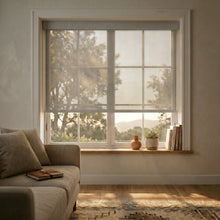 Load image into Gallery viewer, Light Grey Signature solar roller shades installed in a modern living room, featuring a 5% openness factor that filters natural light to protect furniture from UV rays.