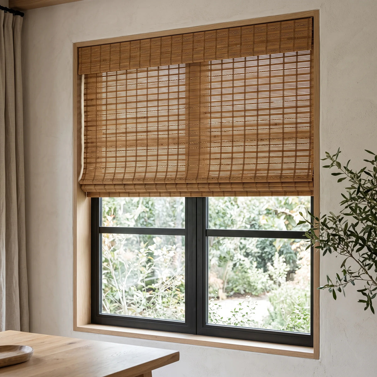 Motorized Woven Wood Shades in warm camel bamboo with a valance, filtering garden light into a dining room with a black-framed window.