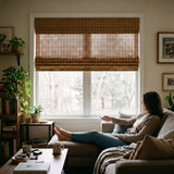 A woman relaxes on the sofa and raises Motorized Woven Wood Shades with the remote, letting soft daylight into the living room.