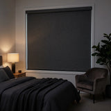 Marine Black Luxe fabric blackout roller shades pulled down in a modern bedroom with a glowing bedside lamp, providing a total dark-sleep environment