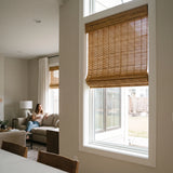 Papara Natural Motorized Woven Wood Shades raised on a tall window while a woman controls them from the sofa with a remote.