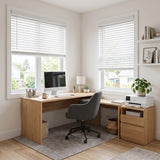 A modern home office setup featuring a light wood L-shaped desk with a gray swivel chair, styled with a computer, lamp, printer, and plants, set against white walls with two windows fitted with white roller blinds.