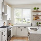 A bright, modern kitchen with white shaker-style cabinets, a white marble countertop, a stainless steel gas range, and a large window with white blinds.