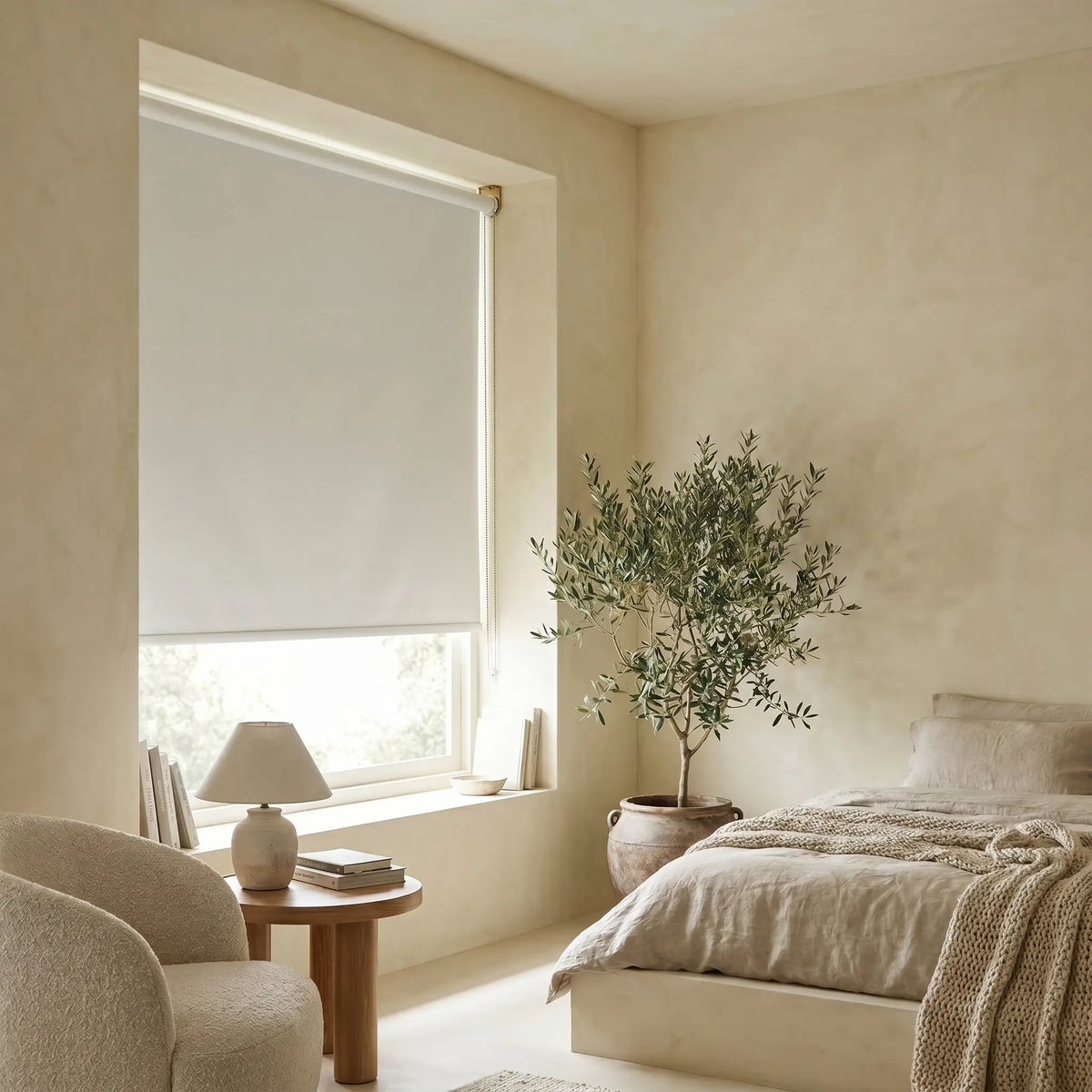 A bright, airy bedroom featuring white Luxe fabric blackout roller shades, styled with a neutral armchair and a large indoor plant